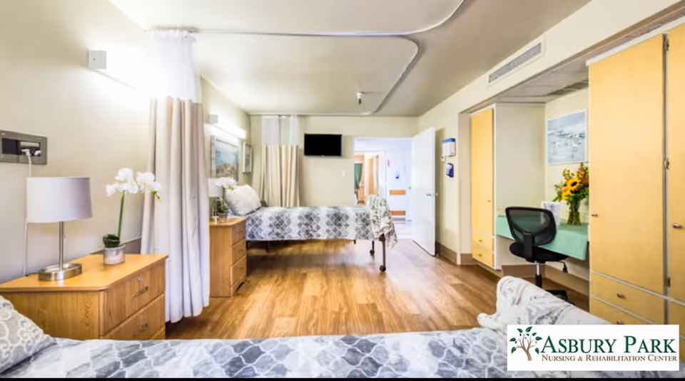 A bright and clean nursing facility room with two beds, wooden furniture including dressers and a desk, a chair, and a wall-mounted TV. The room has light-colored walls, wood flooring, and curtains for privacy between beds. There is a vase with flowers on the desk.