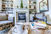 Cozy living room area with two patterned armchairs facing a white fireplace with built-in shelves on either side. The shelves hold decorative items and books. A coffee table in the foreground has a white cup and saucer, a small bowl, and a stack of books. A framed floral painting hangs on the wall to the right.
