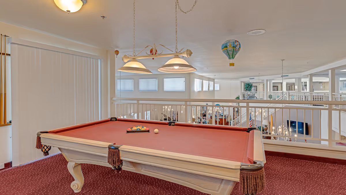 A red felt pool table with billiard balls arranged in a triangle and a cue ball on top, situated in an upper-level recreational area overlooking a spacious, well-lit common area with white railings and hanging light fixtures.