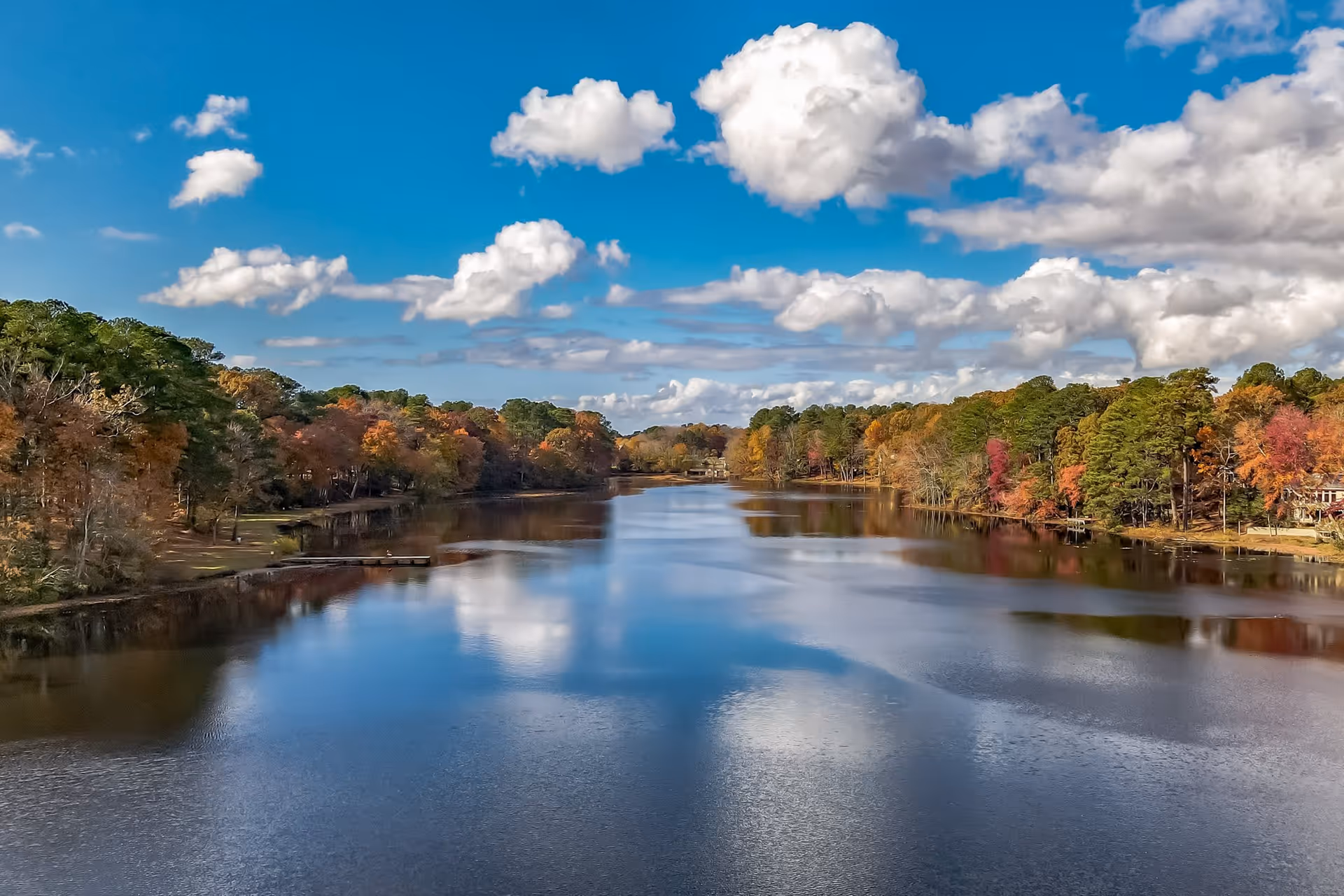 A calm river or lake surrounded by trees with autumn foliage under a blue sky with scattered white clouds.