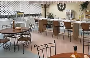 A dining area with round tables and metal chairs in the foreground and a long counter with high chairs in the background. The room has a light-colored floor and walls with lattice paneling and decorative wreaths. Various items are placed on the counter and tables.