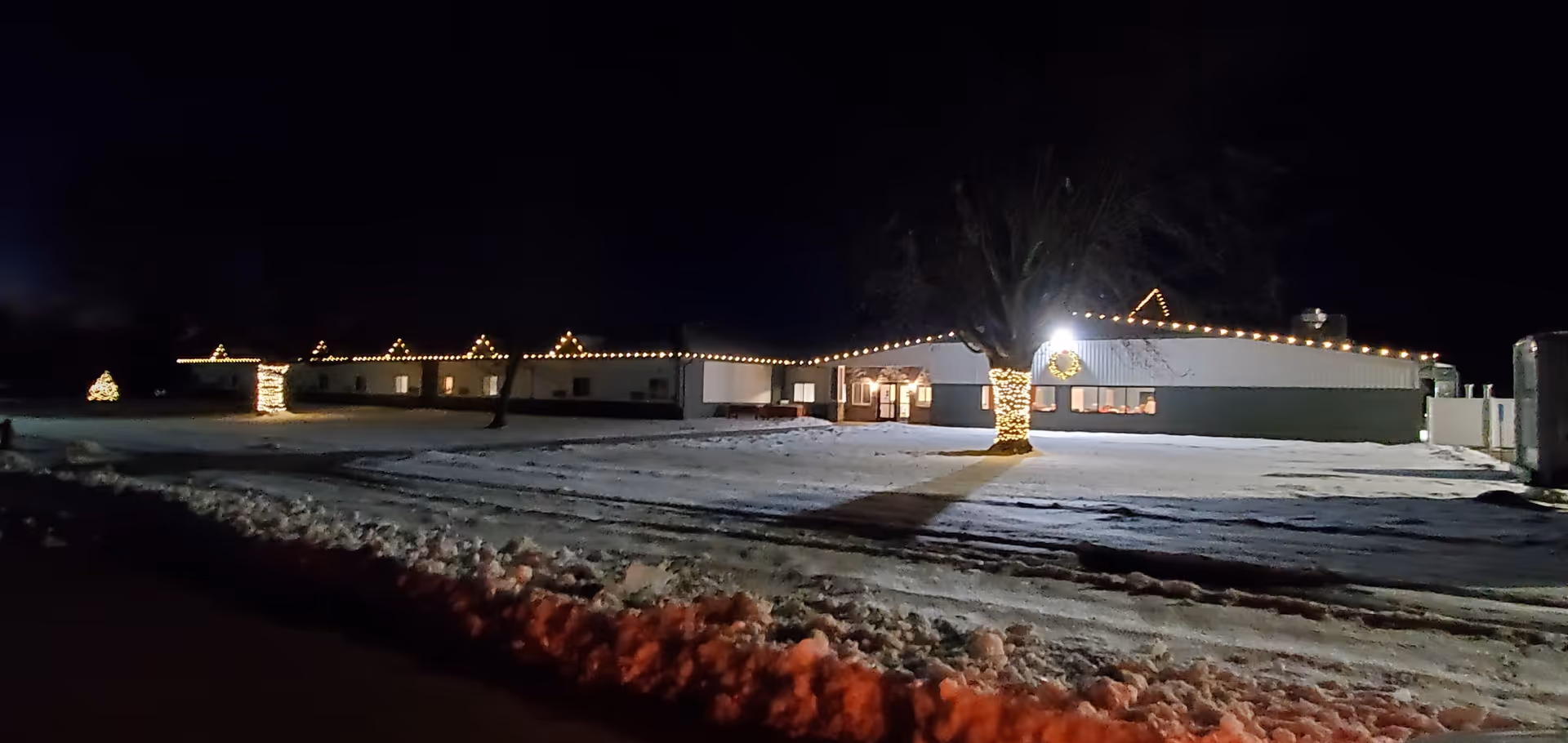 Nighttime exterior view of a single-story building decorated with string lights along the roofline and wrapped around tree trunks. Snow covers the ground and a small decorated tree is visible on the left side of the image.