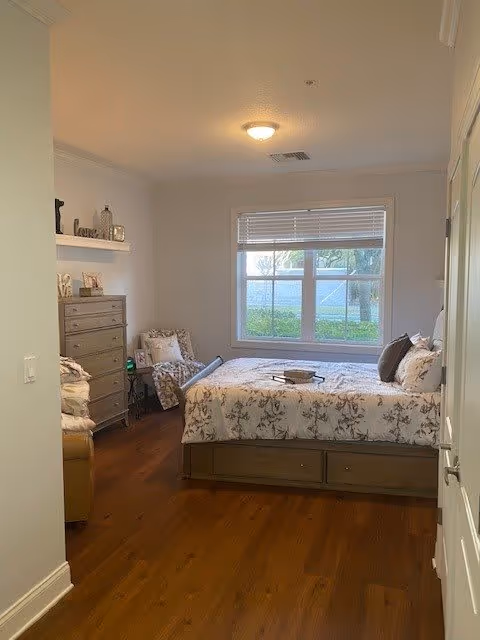 A cozy bedroom with a large window letting in natural light. The room features a bed with floral bedding, a wooden dresser, a cushioned chair with a blanket, and decorative items on a wall shelf. The floor is wooden, and the walls are painted light gray.