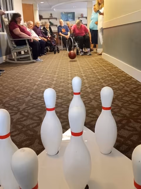 A group of elderly people sitting and watching as a woman in a wheelchair rolls a bowling ball down a carpeted hallway towards a set of white bowling pins with red stripes.