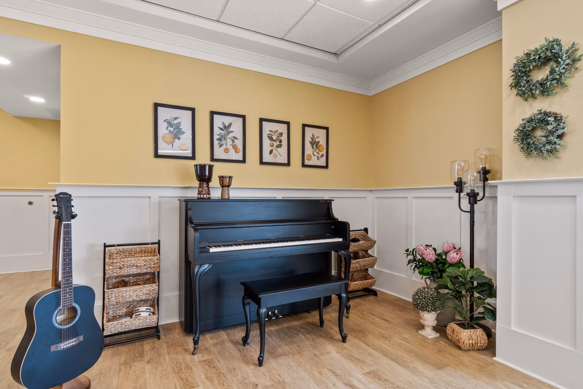 A cozy corner of a senior living facility featuring a black upright piano with a matching bench, a black acoustic guitar resting against the wall, and a wicker basket organizer. The walls are painted yellow with white wainscoting, decorated with four framed botanical prints and two green wreaths. There are also two decorative drums on the piano, a tall candle holder with three candles, and potted plants on the floor.