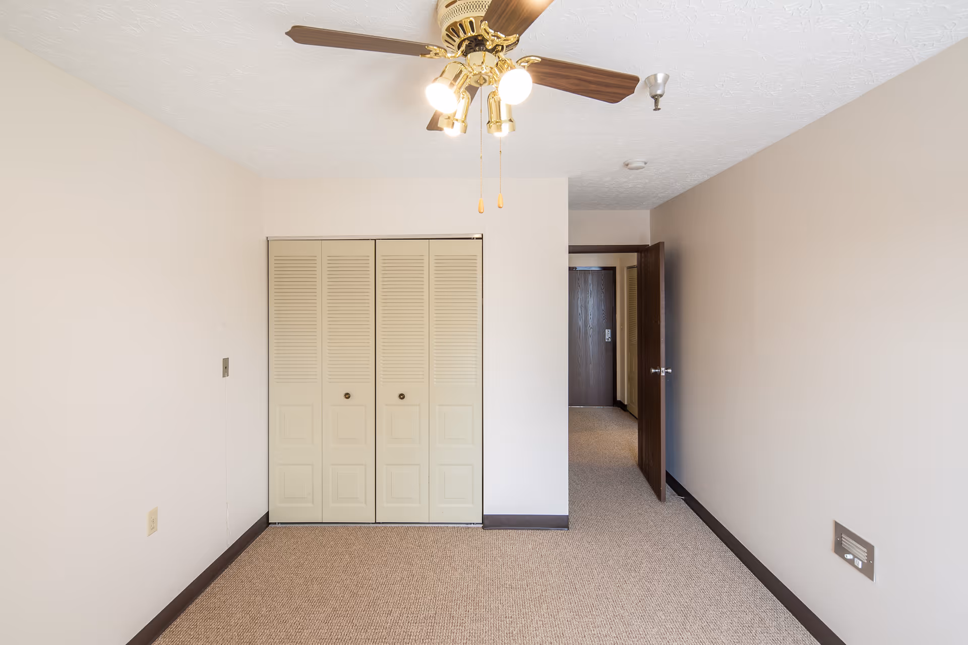 Empty room with beige walls and carpeted floor, featuring a ceiling fan with lights, a closed closet with louvered doors, and an open door leading to a hallway.