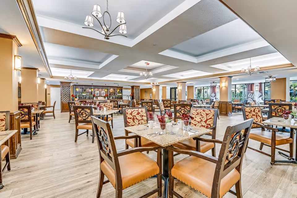 Spacious dining room with multiple square tables and cushioned chairs featuring floral patterned backs. The room has a light wood floor, beige walls with wooden paneling, and several chandeliers hanging from a coffered ceiling. The tables are set with cups, utensils, and folded napkins. Large windows on one side let in natural light.