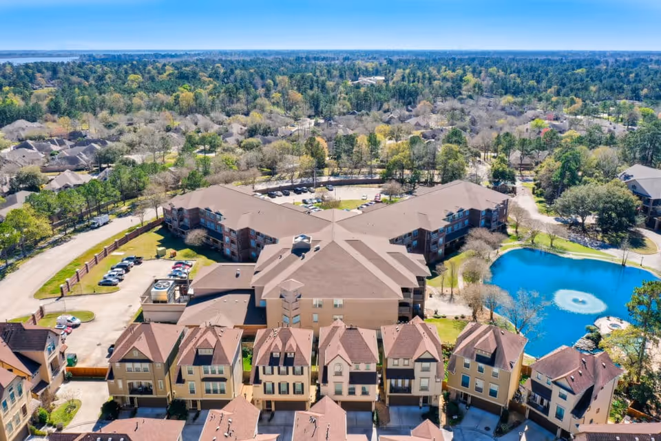 Aerial view of The Terraces at Kingwood Town Center senior living facility showing multiple residential buildings with brown roofs, a parking area, a pond with a fountain, and surrounding trees and greenery under a clear blue sky.