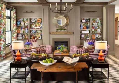 A cozy living room with patterned carpet, two striped armchairs, a dark leather sofa, and a wooden coffee table with a bowl of green apples and an open book. The room features a fireplace with a round mirror above it, built-in shelves filled with decorative items, and two table lamps with orange bases on side tables. Large windows with floral curtains allow natural light to enter.
