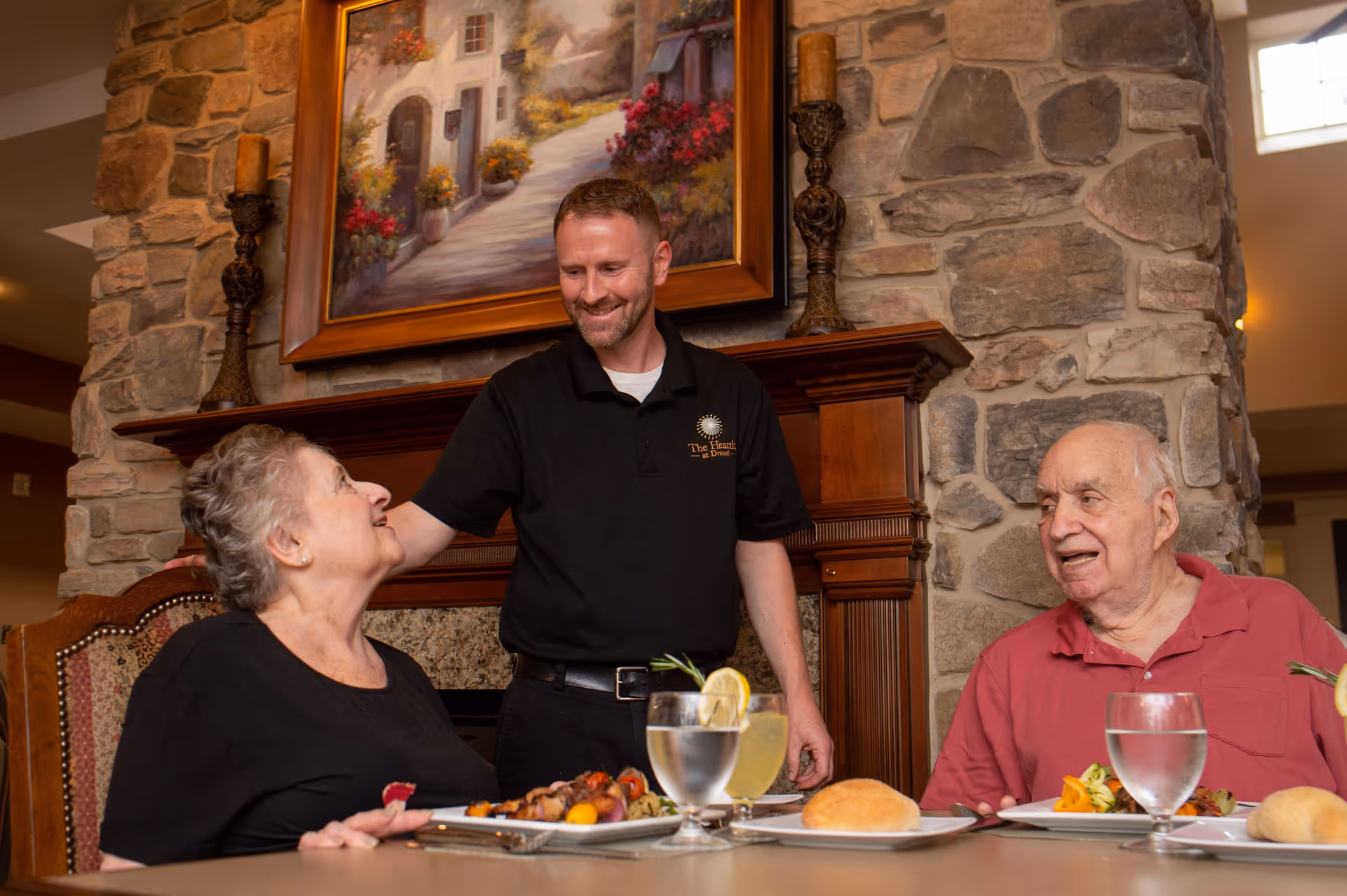 A staff member stands smiling between two elderly residents seated at a dining table with plates, drinks, and a roll in front of a stone fireplace.