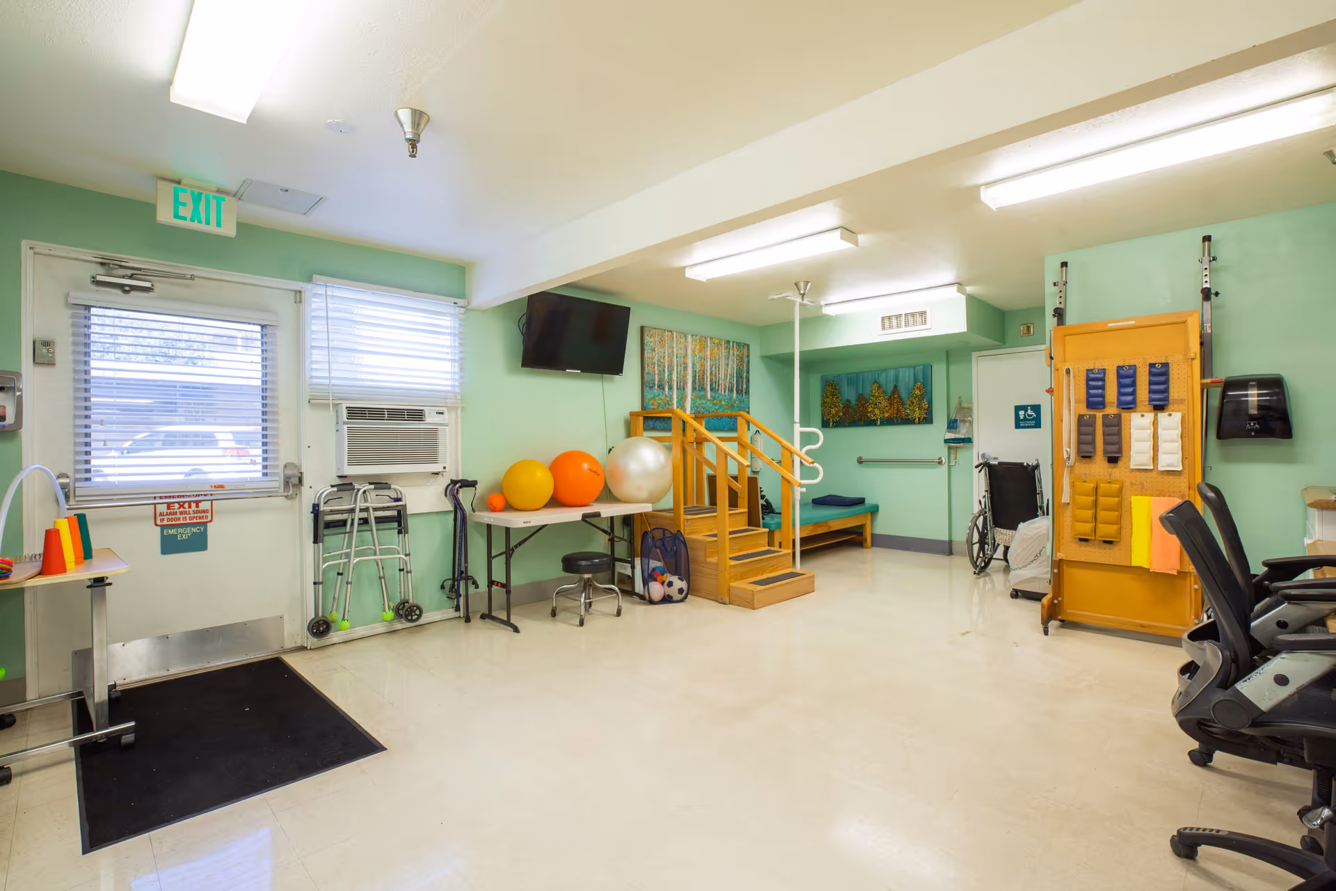 A bright mint-green therapy room with exercise balls, a small wooden practice staircase, walkers, and a wheelchair.