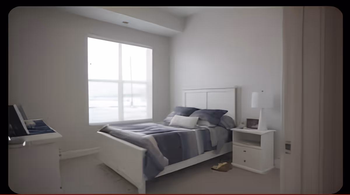 Sunlit simple bedroom with a white bed dressed in blue-gray bedding, a nightstand with a lamp, and a window letting in light.