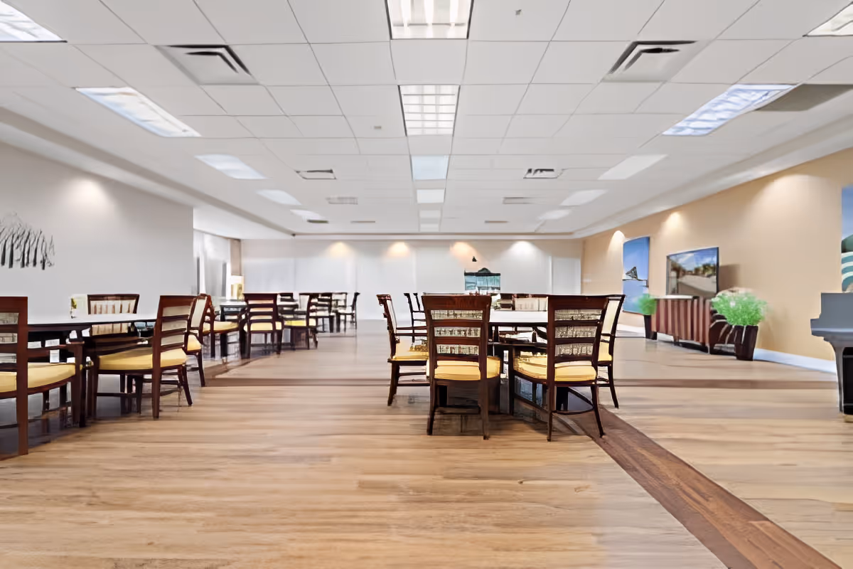 Spacious dining room with multiple wooden tables and chairs arranged neatly on a wood-patterned floor. The room has a white ceiling with recessed lighting and beige walls adorned with paintings and potted plants. A piano is visible on the right side of the room.