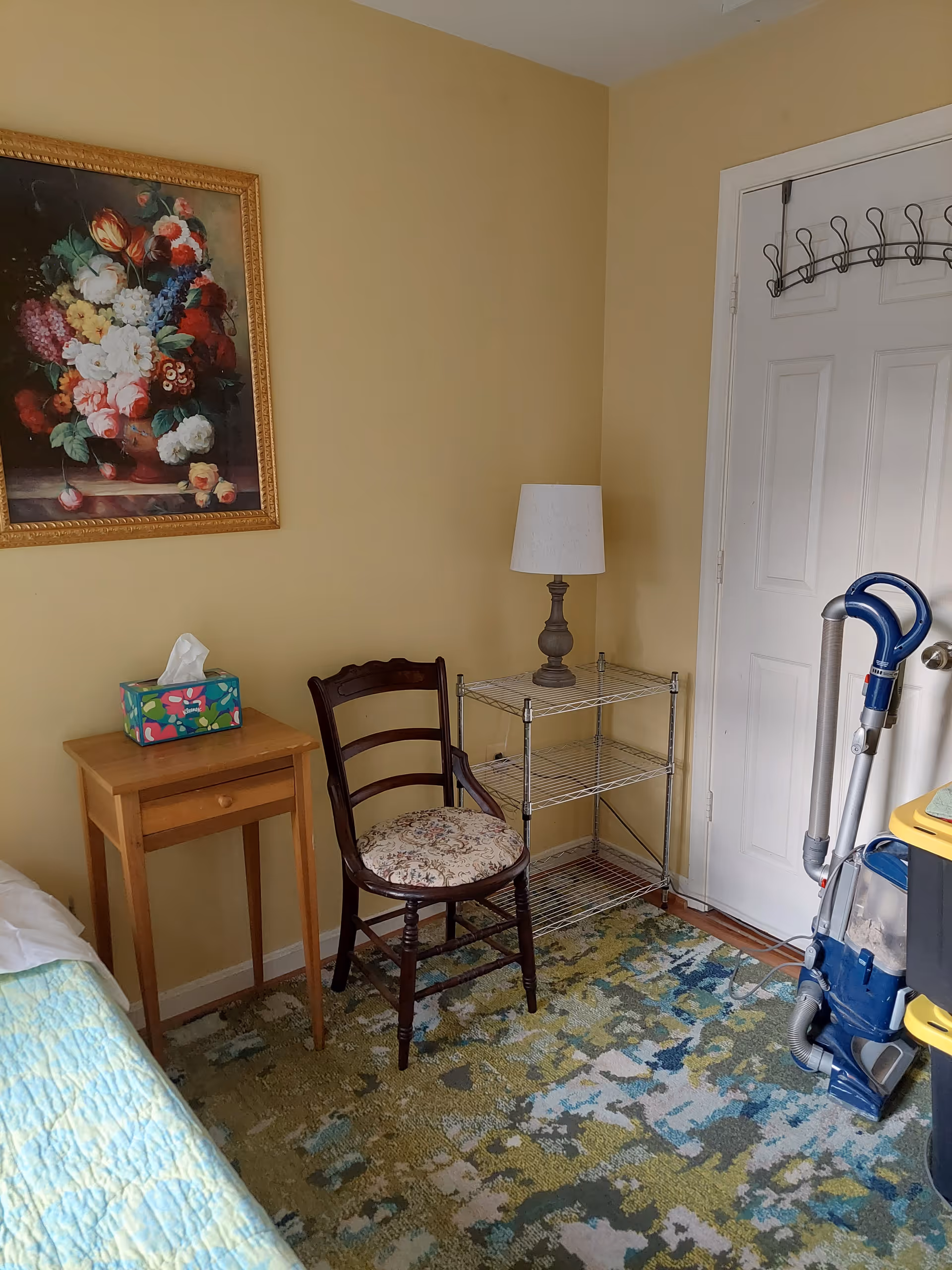 A corner of a bedroom with a wooden chair that has a floral cushion, a small wooden side table with a colorful tissue box on top, a metal shelving unit with a table lamp, a vacuum cleaner, and a white door with a coat rack. The walls are painted yellow and there is a framed painting of flowers hanging on the wall. The floor has a patterned carpet with green, blue, and beige colors.