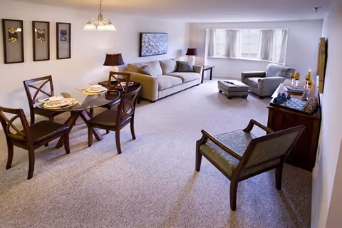 A spacious living room and dining area with beige carpet. The dining area has a round glass table set with four wooden chairs and place settings. The living room features a beige sofa, an armchair with an ottoman, a wooden chair, two side tables with lamps, a wall-mounted painting, and a large window with white curtains letting in natural light. A wooden cabinet with decorative items is also visible.