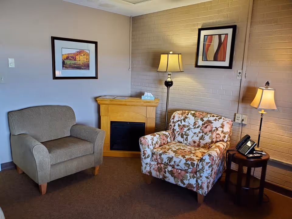 Cozy sitting area with two armchairs, a small mantel-style electric fireplace, side table with a phone, and lamps against a brick wall.