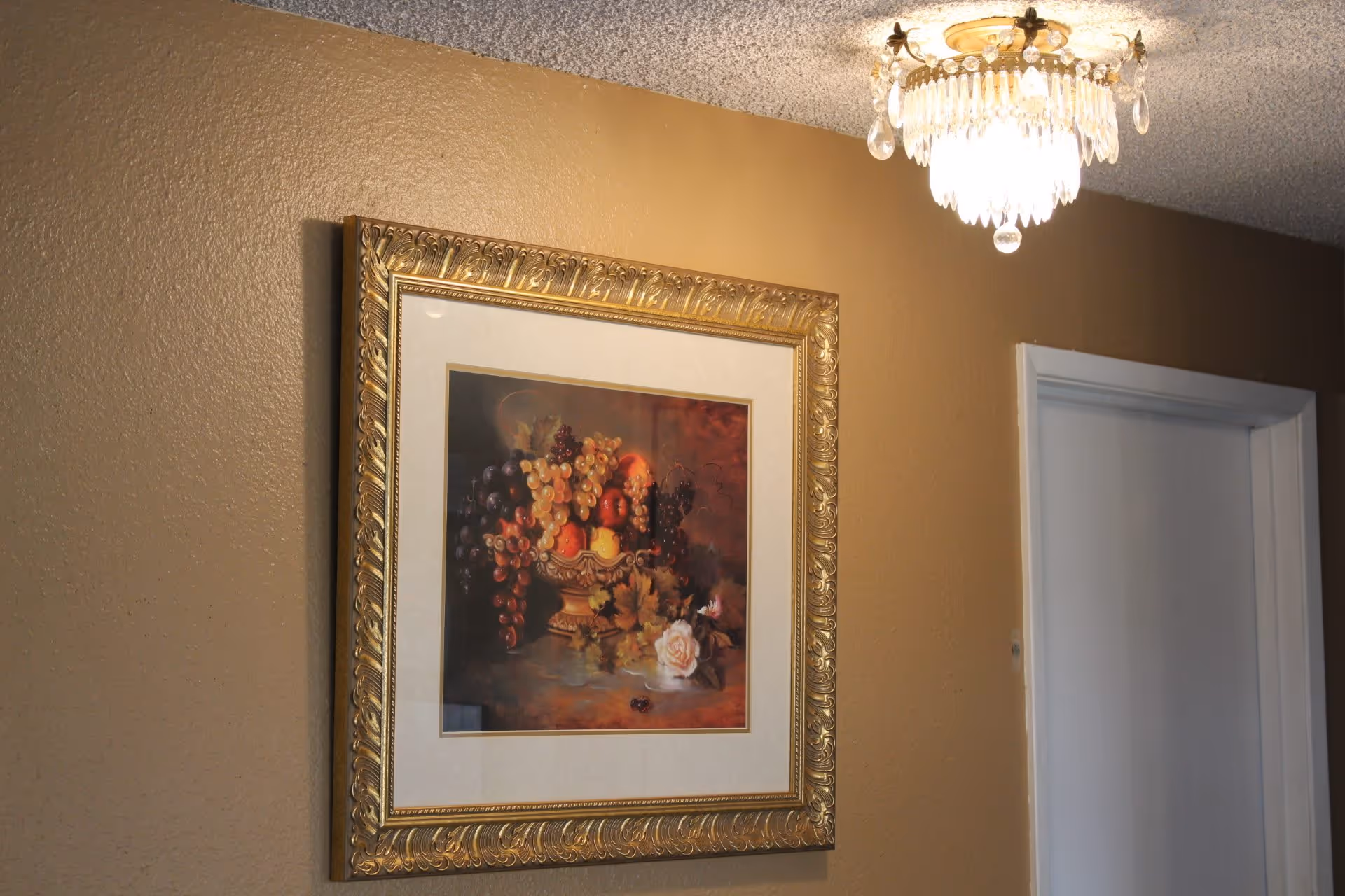 A framed painting of a fruit basket with grapes, apples, and a rose, hanging on a beige textured wall next to a white door. A decorative chandelier with hanging crystals is mounted on the ceiling above.