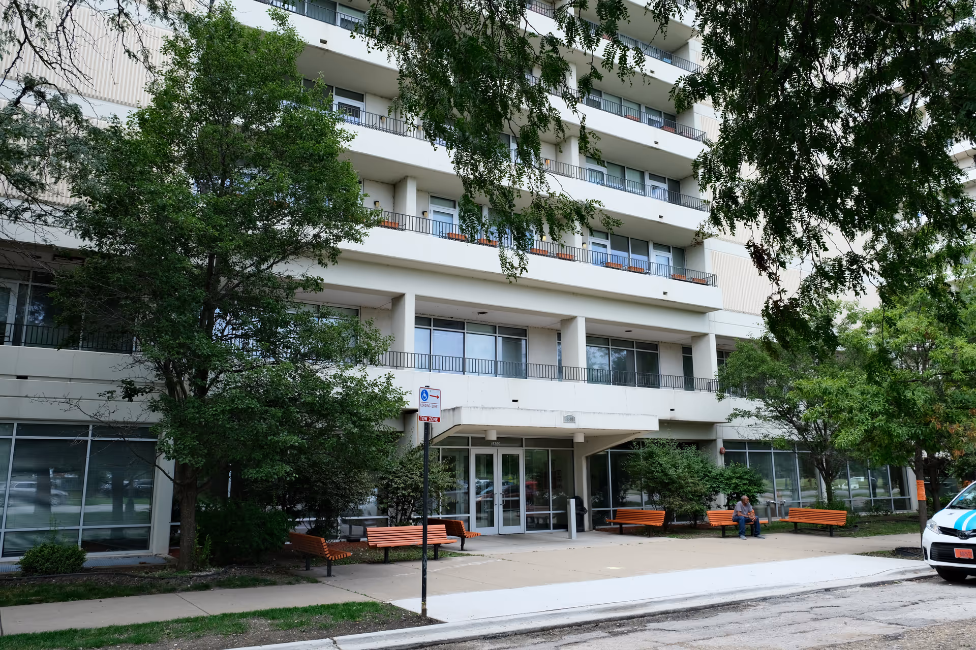 Exterior view of a multi-story senior living facility named Pioneer Gardens with balconies, trees, benches, and a person sitting on one of the benches near the entrance.