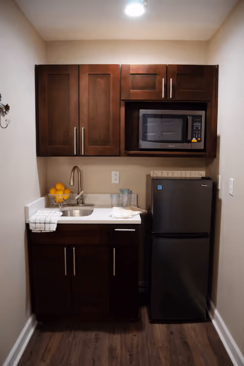 Small kitchenette with dark wooden cabinets, a stainless steel sink, a bowl of oranges, a microwave, a mini refrigerator, and a towel hanging on the counter edge, all set against beige walls and wood flooring.