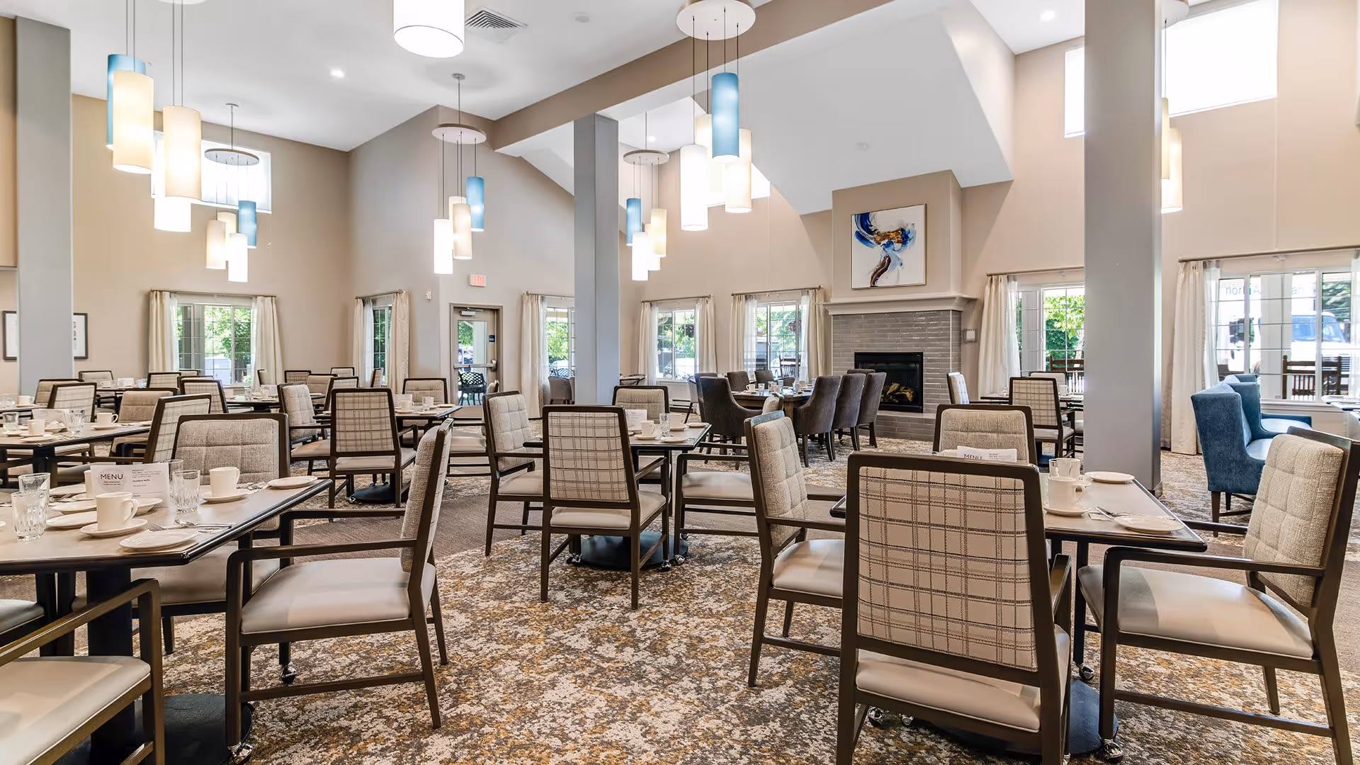 A spacious dining room with multiple tables and chairs arranged neatly. The room features high ceilings with modern pendant lights hanging down, large windows with beige curtains allowing natural light to fill the space, and a fireplace with artwork above it. The tables are set with white cups, glasses, and menus, creating a welcoming atmosphere.