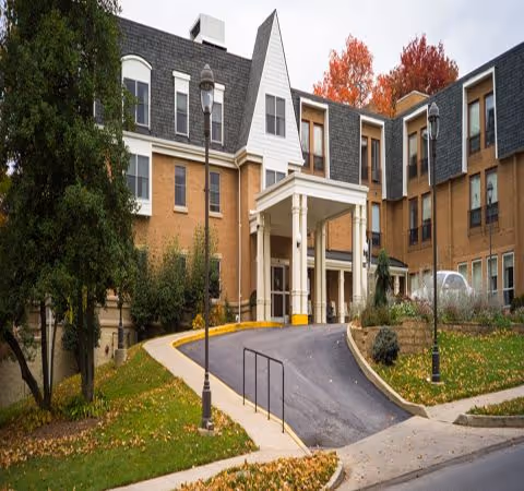 Exterior view of a multi-story senior living facility building with a covered entrance driveway, surrounded by trees with autumn foliage and street lamps along the driveway.