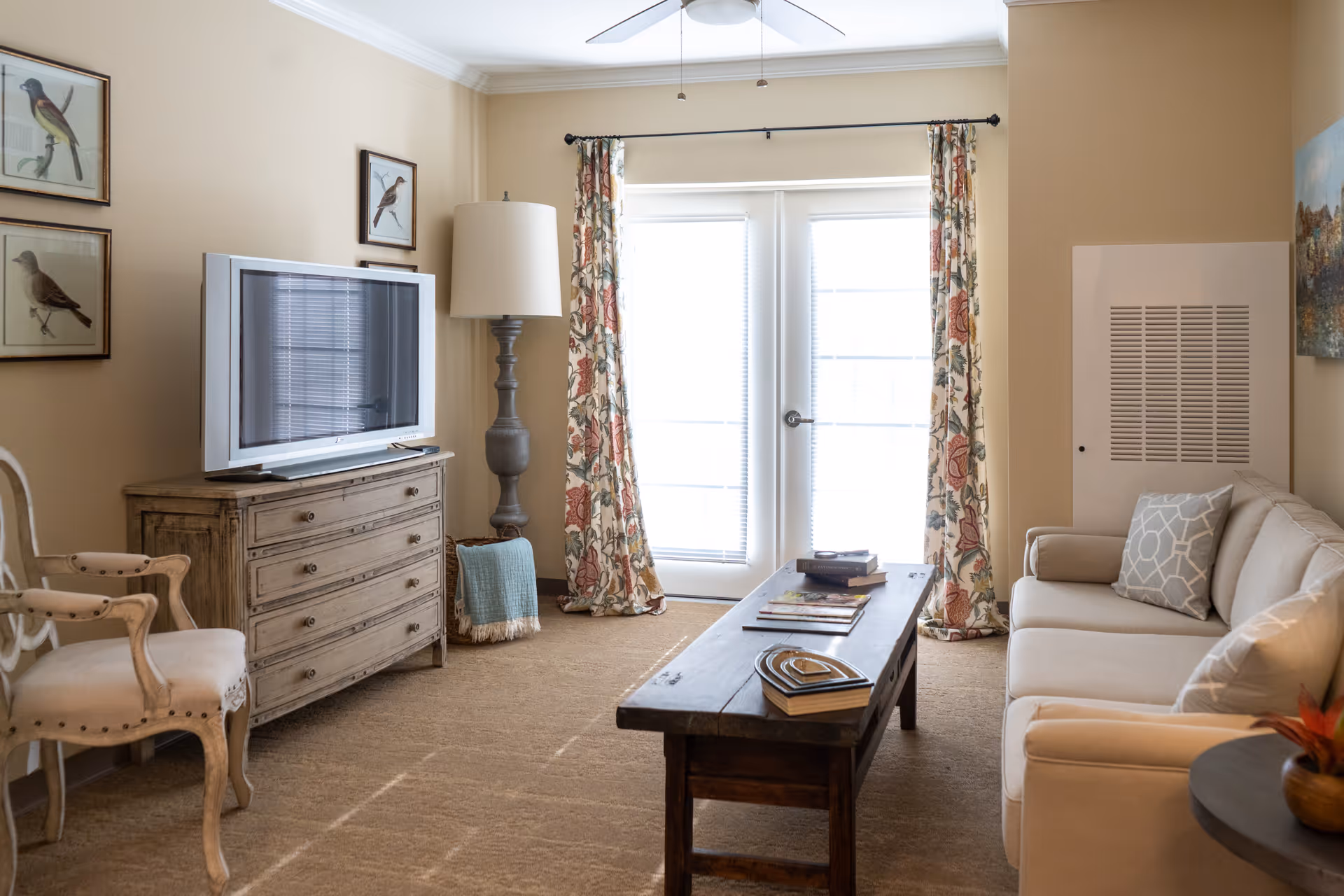 A cozy living room with a beige sofa adorned with patterned pillows, a wooden coffee table with books and decorative items, a wooden dresser with a flat-screen TV on top, a tall floor lamp, and floral curtains framing glass doors that let in natural light. There are framed bird pictures on the wall and a beige armchair with wooden arms.