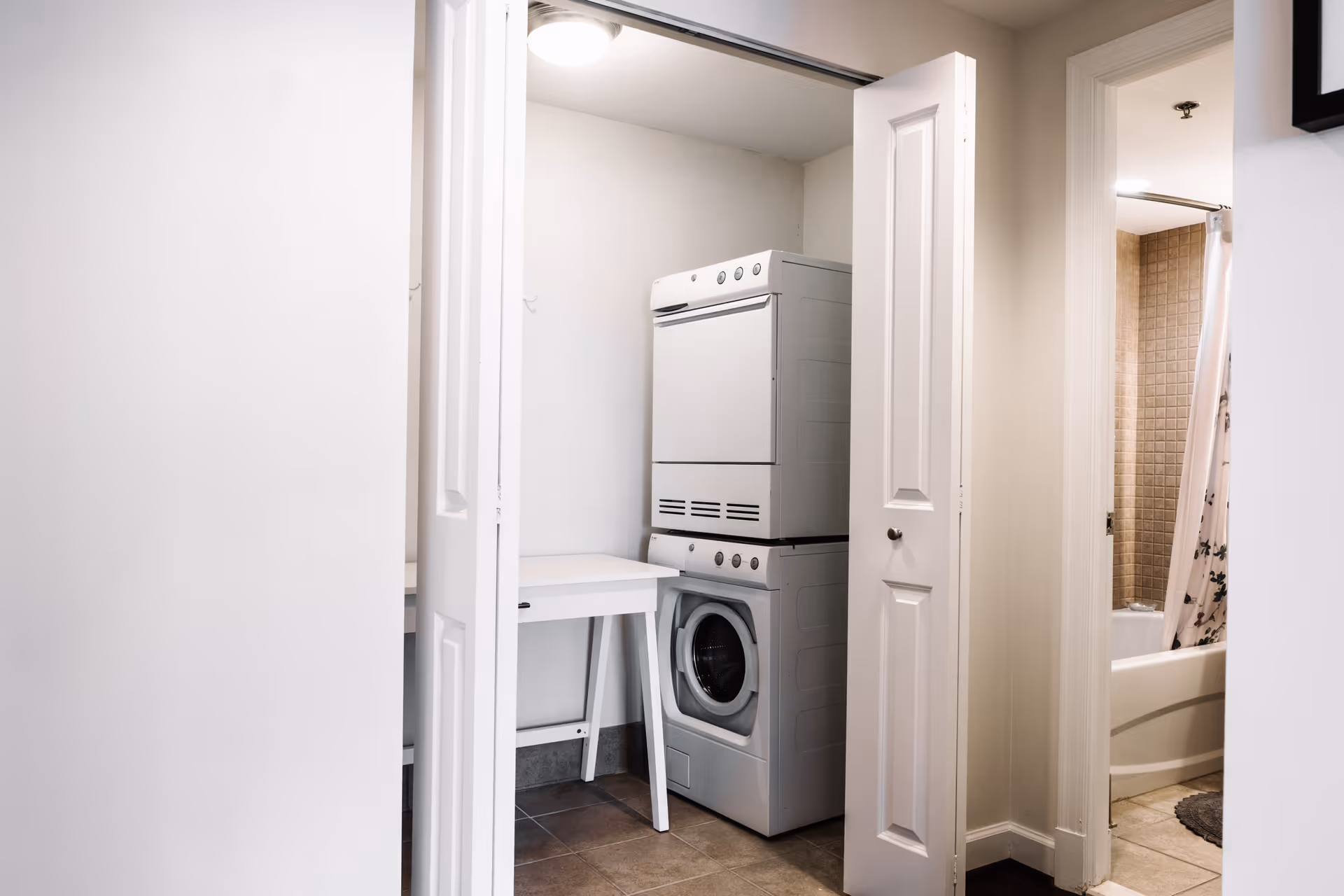 A small laundry area with a stacked washer and dryer inside a closet with white bi-fold doors. To the right, there is a bathroom with a bathtub and a shower curtain visible.