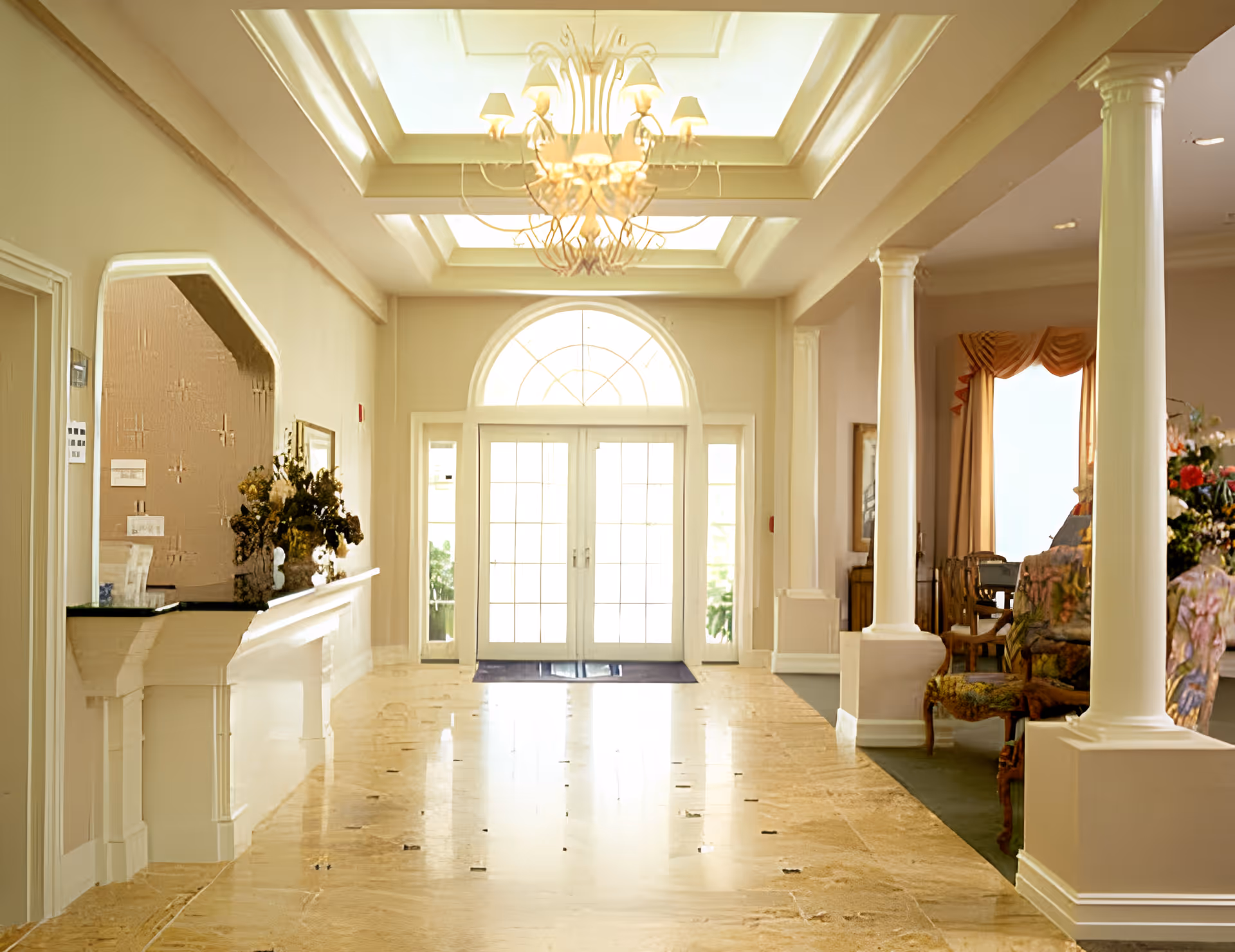 Bright and elegant hallway in Mathison Retirement Community featuring a polished marble floor, white columns, a decorative chandelier, and a large arched glass door letting in natural light. There is a seating area with floral upholstered chairs and a table with flowers on the right side.