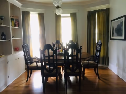A dining room with a wooden oval dining table surrounded by six dark wooden chairs with cushions. The room has wooden flooring, three tall windows with green curtains, a built-in white cabinet with shelves on the left, and a framed picture on the right wall. A chandelier hangs from the ceiling above the table.