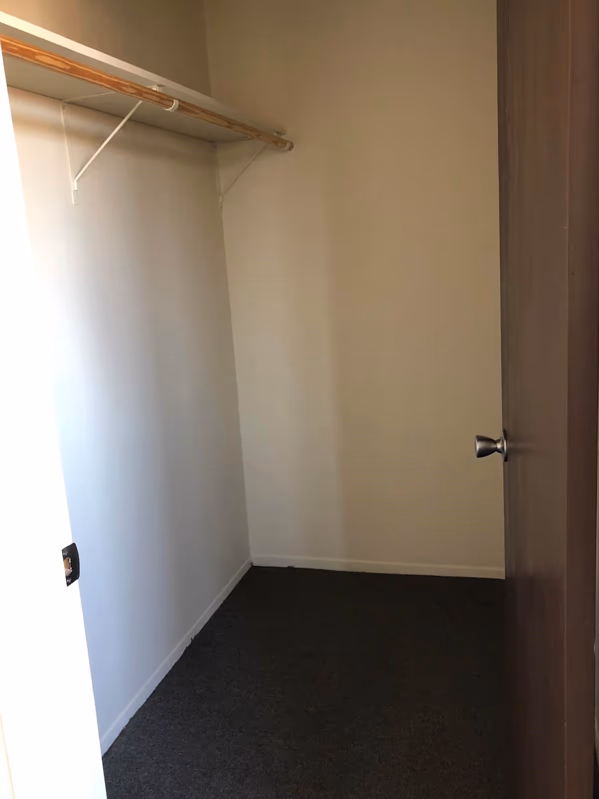 Empty walk-in closet with a wooden hanging rod supported by white brackets, beige walls, dark carpeted floor, and a partially open wooden door with a silver knob.