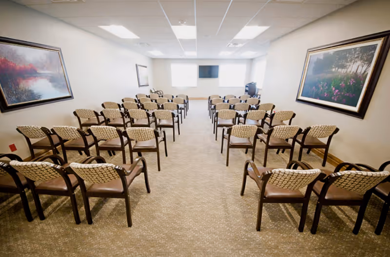 Rows of chairs arranged classroom-style facing a wall-mounted TV in a spacious meeting/activity room with framed artwork on the walls.