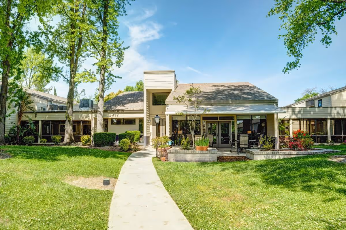 Exterior view of a single-story building with a sloped roof surrounded by green grass, trees, and shrubs under a clear blue sky. A concrete pathway leads to the entrance, which has large windows and a covered patio area with outdoor seating.