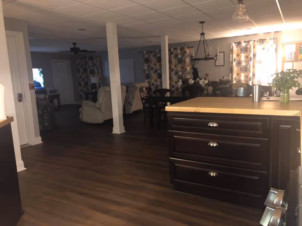 Open communal interior showing a kitchen island in the foreground, a dining table and chairs, and recliner seating with a TV in the background.