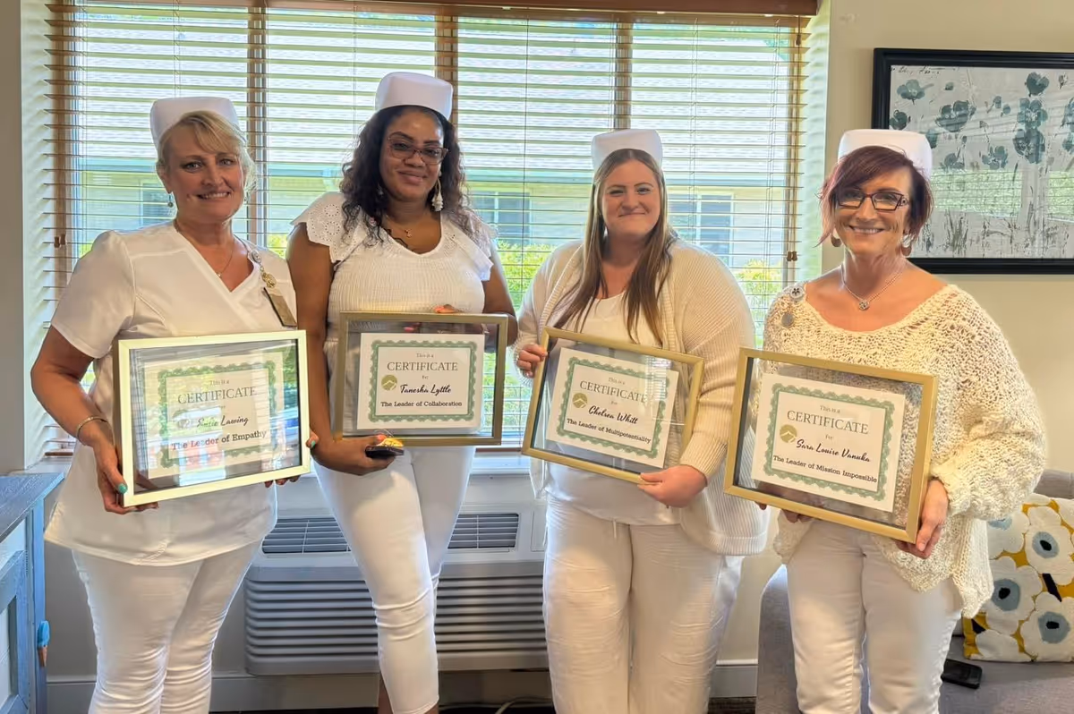 Four women dressed in white nursing uniforms and caps standing indoors in front of a window with blinds, each holding a framed certificate. They are smiling and posing for the photo in a room with a couch and decorative pillows.