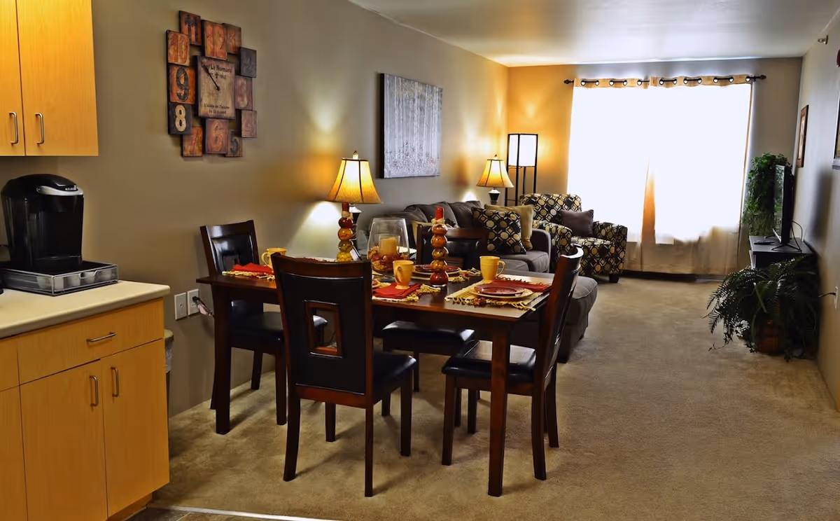 Open living and dining area in a senior living apartment with a set dining table, sofas, lamps, and a TV by a bright window.