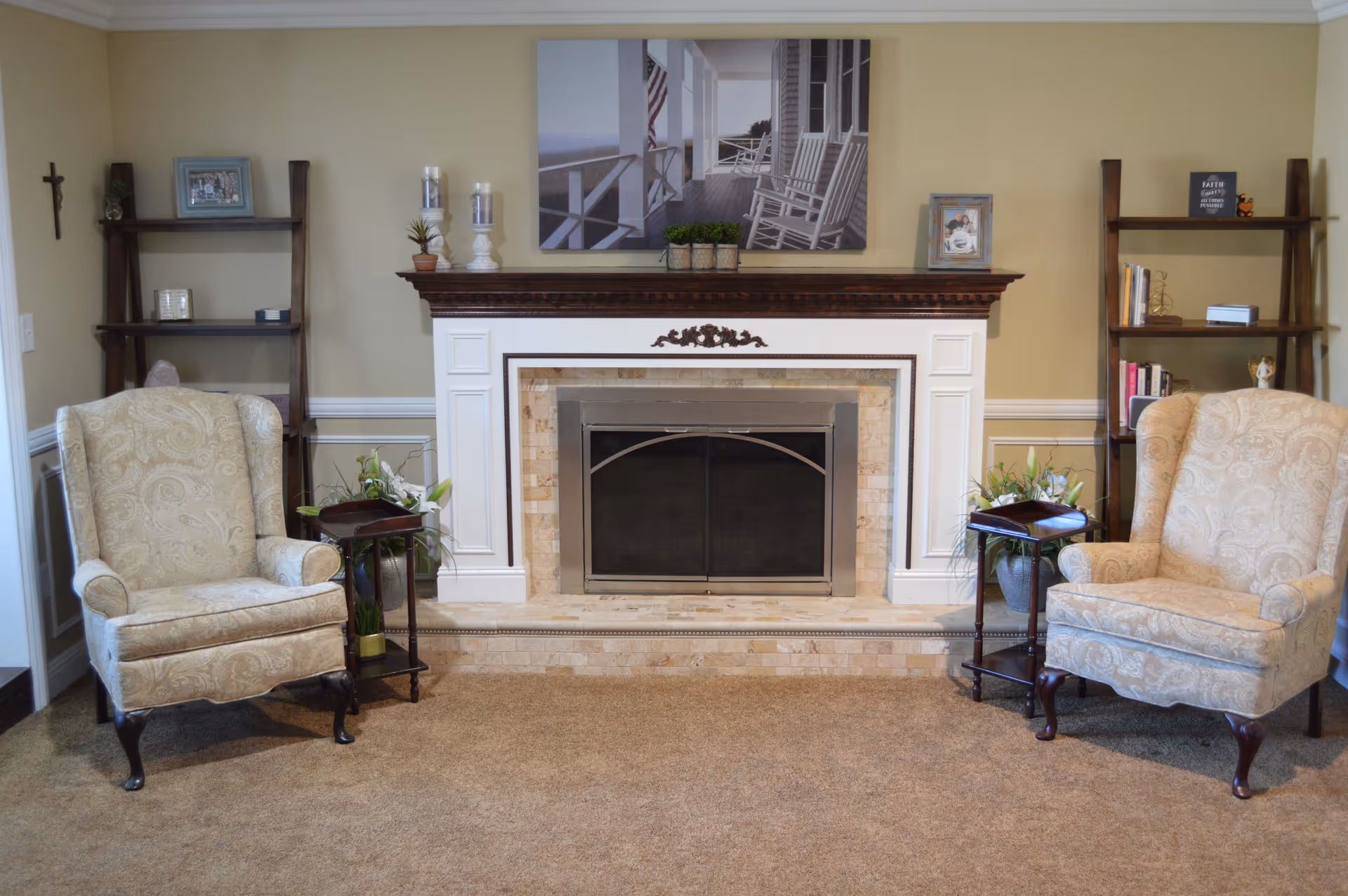 A cozy living room area with two beige patterned armchairs on either side of a white fireplace with a dark wooden mantel. The fireplace has a metal screen and is surrounded by light-colored tiles. On the mantel are decorative items including candles, small plants, and framed pictures. Behind each armchair is a wooden shelving unit with books and decorative items. The walls are painted beige with white trim, and the floor is carpeted in a light brown color.