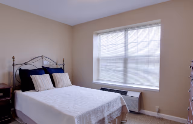 A bedroom with a metal framed bed covered with a white quilt and four pillows, two dark blue and two beige. There is a wooden nightstand with a drawer on the left side of the bed and a wooden dresser on the right side of the image. A large window with closed blinds is on the wall behind the bed, and a heating/cooling unit is beneath the window. The walls are beige and the carpet is light-colored.