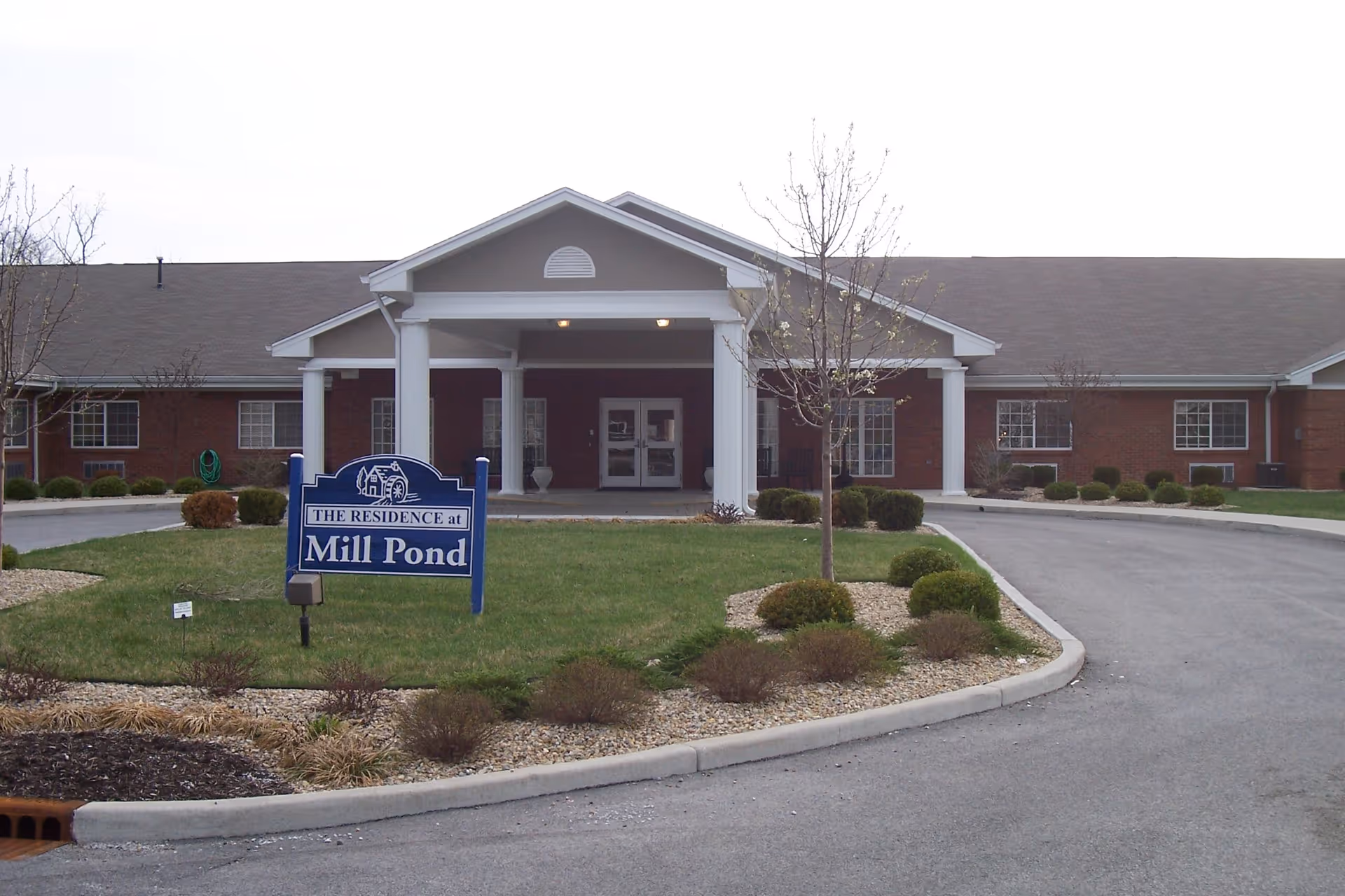 Front entrance of Mill Pond Health Campus, a single-story brick building with white columns, a covered portico, driveway, and a lawn sign reading "Mill Pond".
