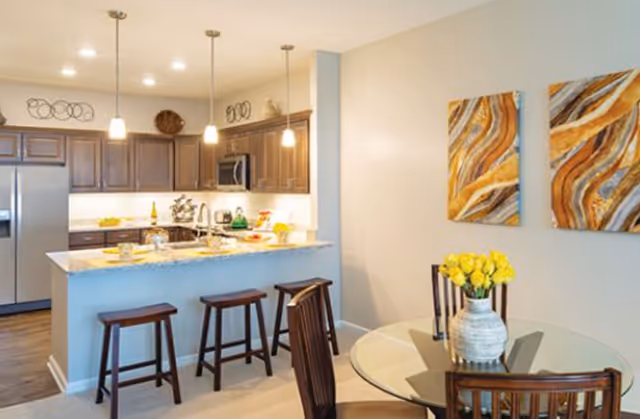 A modern kitchen and dining area in a senior living facility. The kitchen features dark wood cabinets, stainless steel refrigerator and microwave, a granite countertop island with three wooden bar stools, and pendant lights hanging above. The dining area has a round glass table with wooden chairs and a vase of yellow flowers. Two abstract paintings with brown, white, and gold tones hang on the wall.