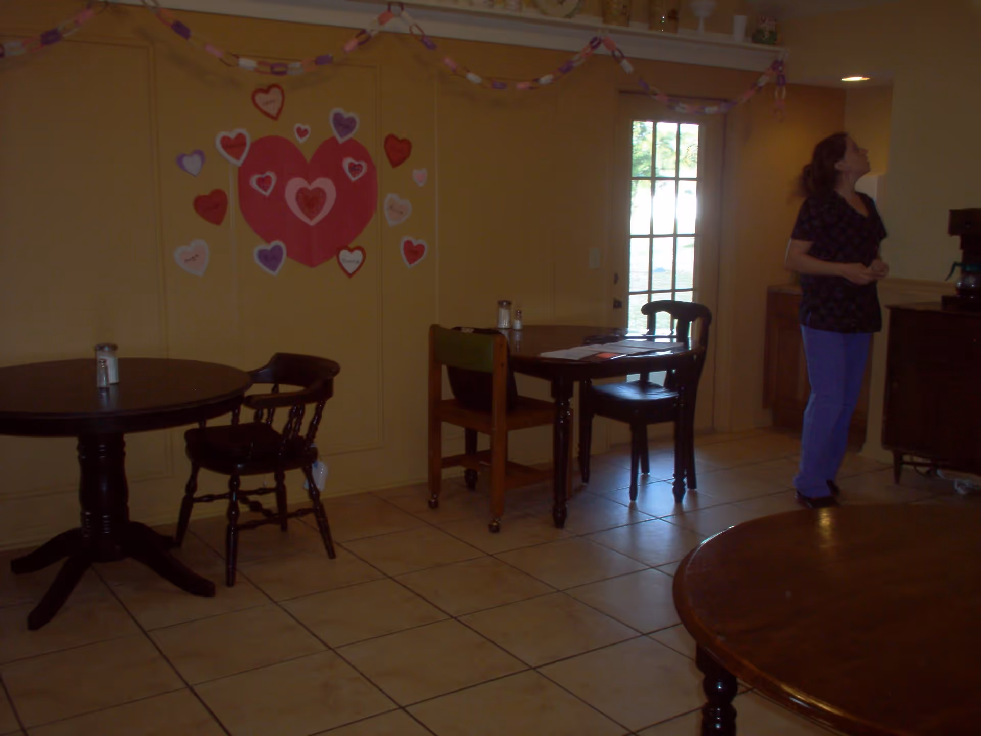 Interior room with several wooden tables and chairs on a tiled floor. A woman in scrubs stands near a counter on the right side of the image. The wall has heart-shaped decorations and paper chains hanging as decorations. A door with glass panels is visible in the background.