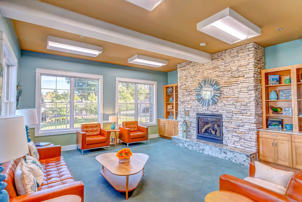 A bright and cozy living room with large windows showing a green outdoor area. The room features a stone fireplace with a decorative round mirror above it, wooden built-in shelves on either side filled with books and decorative items, and orange leather seating including a sofa and armchairs. A wooden coffee table with a small flower arrangement sits in the center on a blue carpeted floor. The ceiling is painted a warm tan color with modern rectangular light fixtures.