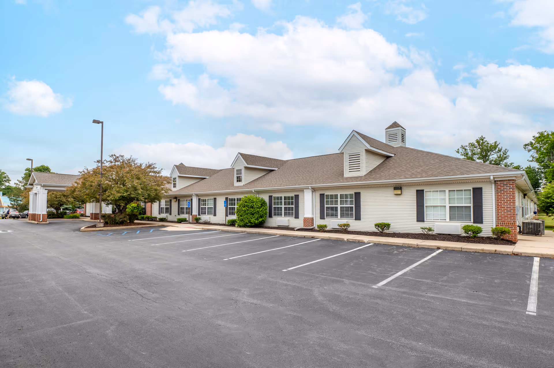 Exterior view of a single-story senior living facility building with a large parking lot in front. The building has a beige siding with brick accents, multiple windows with black shutters, and a covered entrance area. The sky is partly cloudy with blue patches visible.