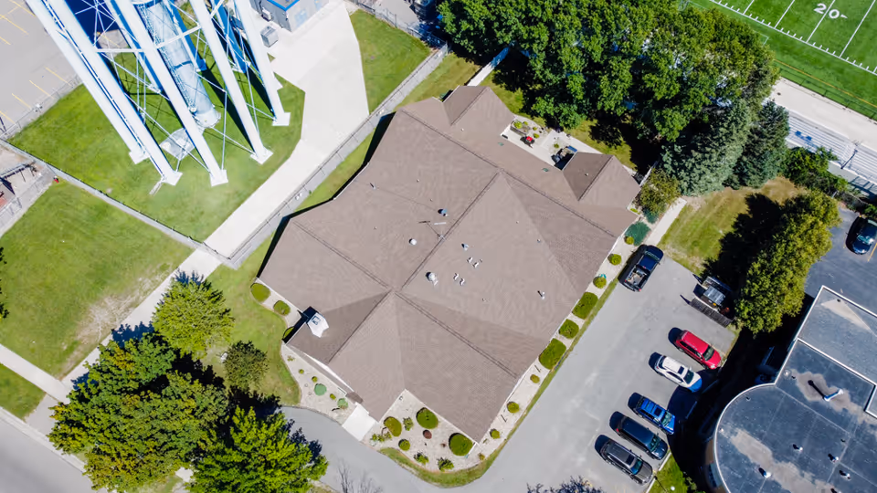 Aerial view of a building with a brown roof surrounded by parking spaces, trees, and a grassy area with a water tower nearby.