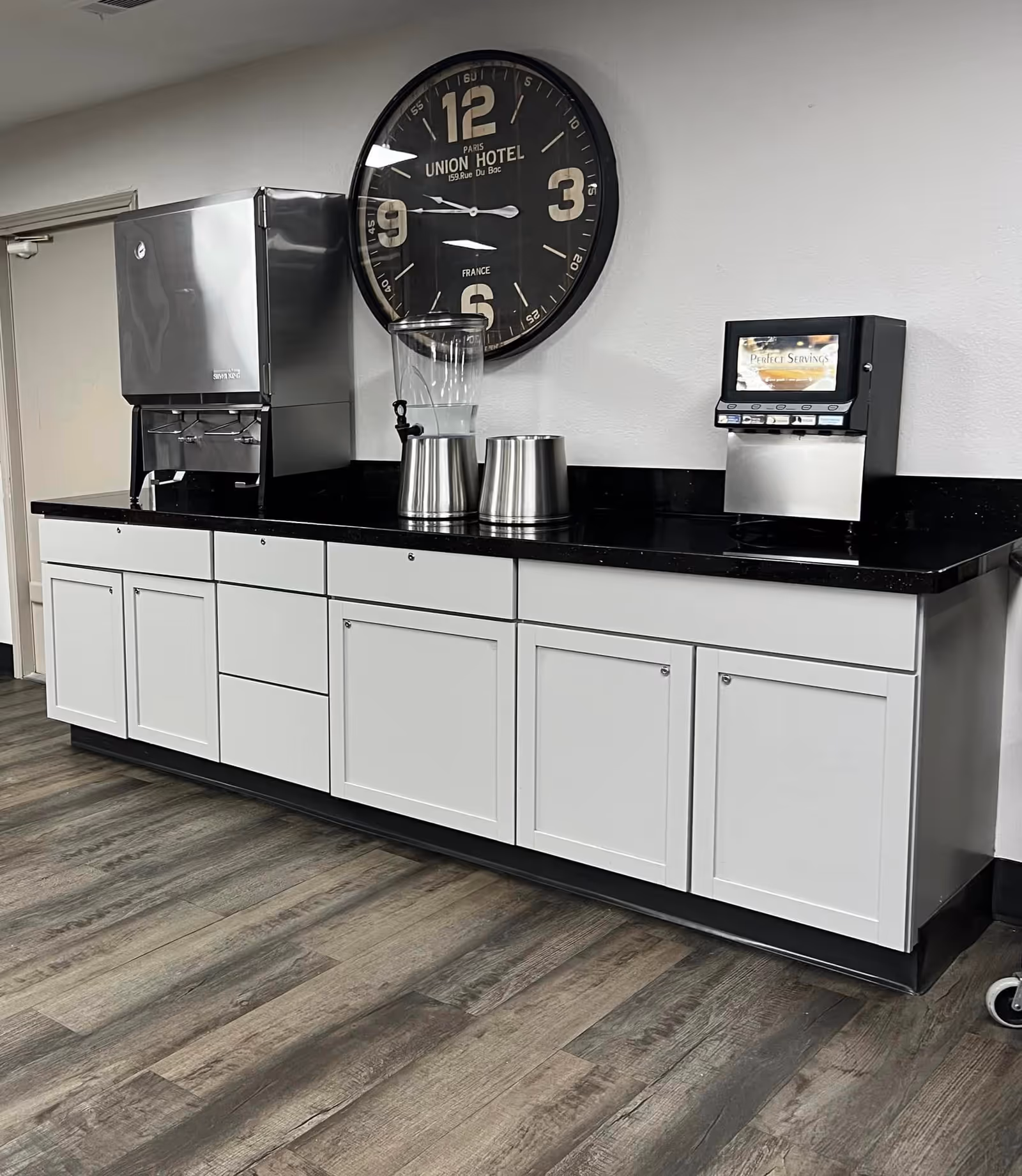 A beverage station with white cabinets, a black countertop, drink dispensers, and a large round wall clock above.