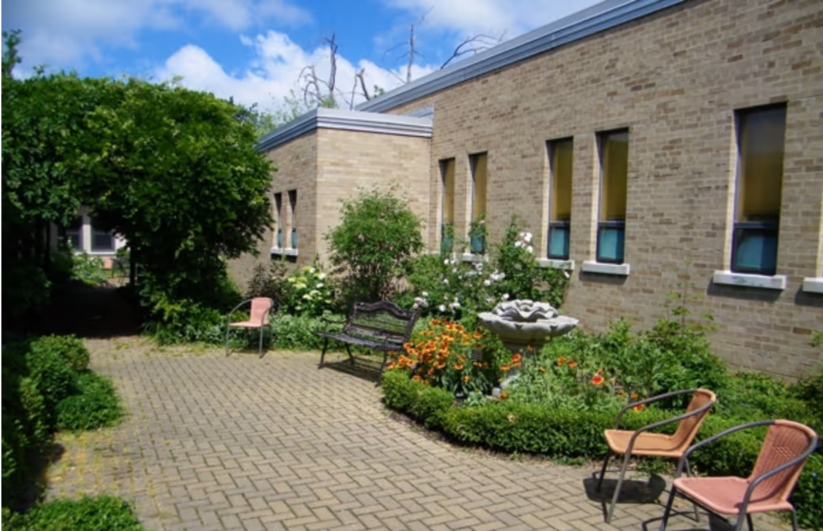 Brick courtyard beside a senior living building with a fountain, benches, chairs, and flowering plants under a blue sky.