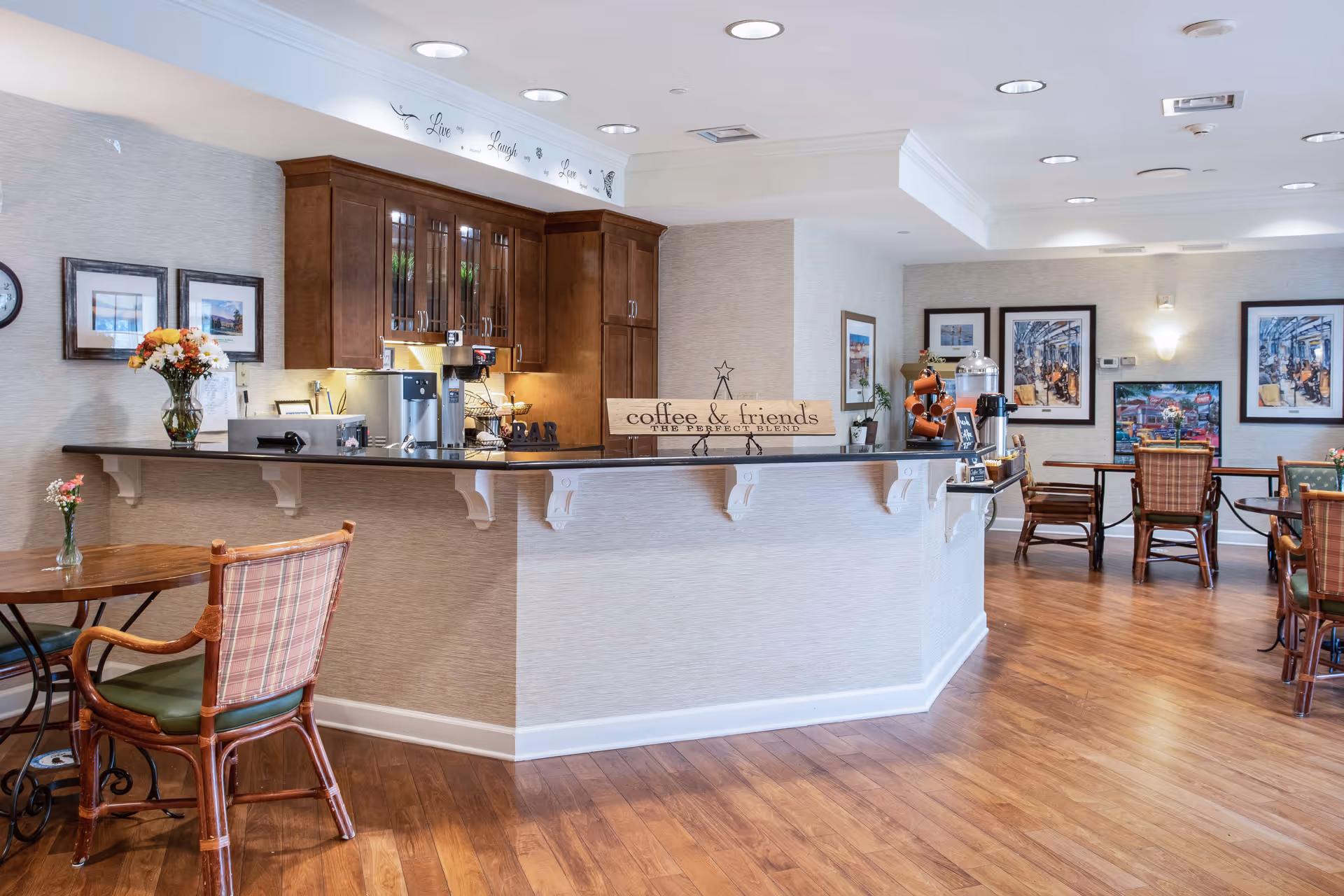 A cozy coffee bar area in a senior living facility with wooden flooring, a counter with coffee machines and a sign that reads 'coffee & friends the perfect blend'. There are tables and chairs with floral arrangements, framed pictures on the walls, and soft lighting from ceiling fixtures.