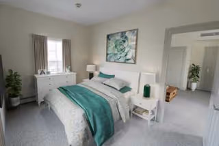 A bright and clean bedroom with a neatly made bed featuring white and teal bedding. There is a white dresser with a lamp and decorative items near a window with beige curtains. A large floral artwork hangs above the bed. The room has light-colored walls and carpeted flooring. A doorway leads to another room with a plant visible inside.