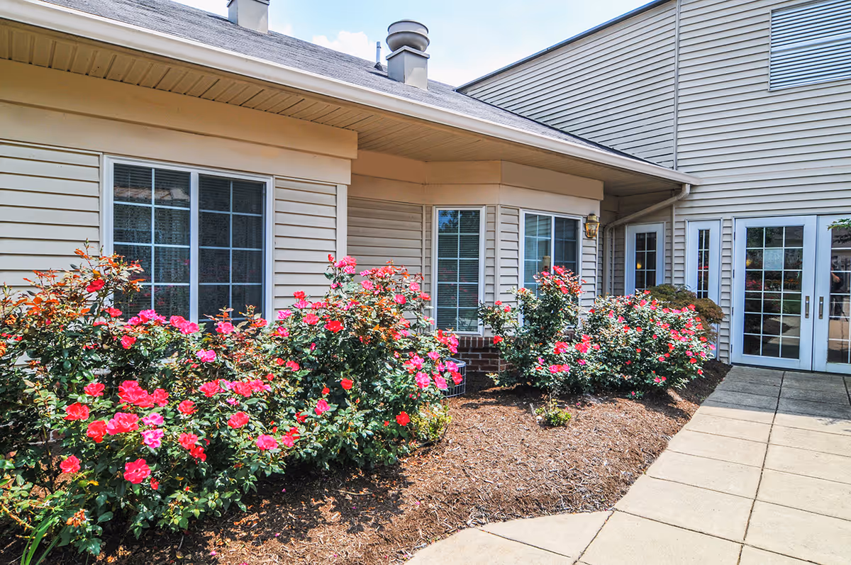 Exterior view of a building with beige siding, multiple windows, and a glass door entrance. In front of the building, there is a garden bed with blooming pink and red flowers and a paved walkway.