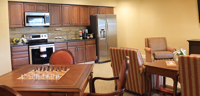 A cozy room featuring a kitchen area with wooden cabinets, a stainless steel refrigerator, microwave, and stove. In the foreground, there is a wooden table with a chessboard and chess pieces set up, surrounded by chairs. To the right, there are comfortable armchairs and a small wooden table with a book and flowers on it.