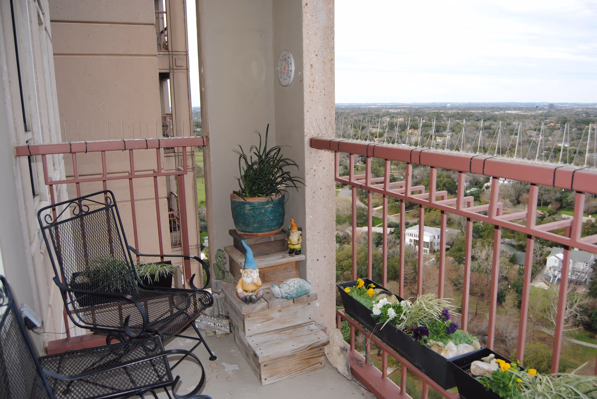 A balcony with two black metal chairs, a potted plant on a wooden stand, garden gnome decorations, and flower boxes attached to the red railing overlooking a green landscape and distant buildings.